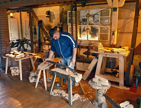 Ein Mann schnitzt mit traditionellem Werkzeug. Im Hintergrund sind weitere Geräte und Holzpantoffeln in verschiedenen Arbeitsstadien zu sehen.