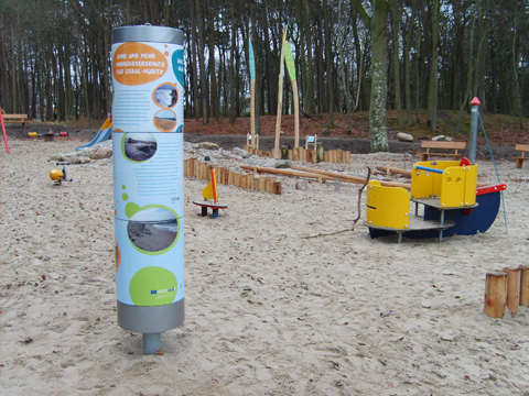 Spielplatz aus Sand mit Geräten aus Holz und einer runden Infosäule