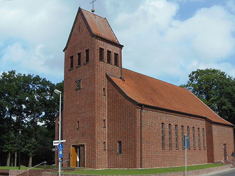 Kirche mit einem Turm. Gebäude, Umgebungsmauer und Treppenaufgang aus rotem Klinker, darum eine Rasenfläche, dahinter alter Baumbestand