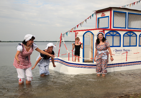 drei Damen im Wasser neben einem hölzernen Boot mit Kabine, auf dem ein Junge steht. Alle tragen historische Badekleidung
